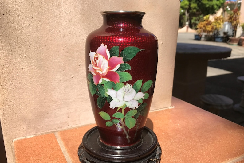 May include: A maroon vase with a floral design. The vase features a red and white rose design with green leaves. The vase is on a black stand. The vase has a silver rim.