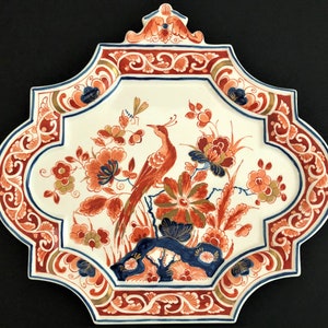 May include: Decorative ceramic plate with a bird and floral design. The plate features a white background with a red, blue, and gold border. The bird is red, and the flowers are orange and blue.