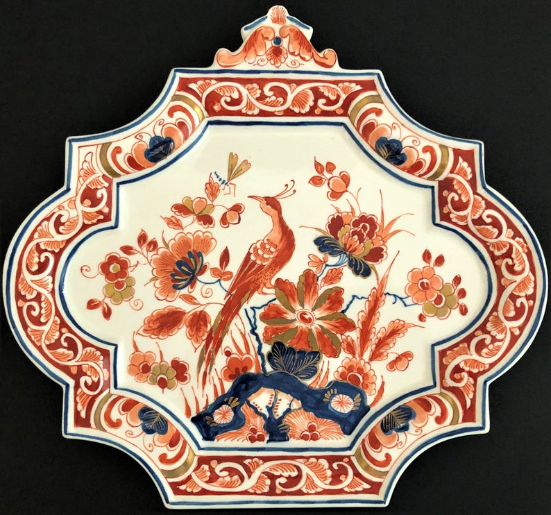 May include: Decorative ceramic plate with a bird and floral design. The plate features a white background with a red, blue, and gold border. The bird is red, and the flowers are orange and blue.