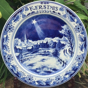 K&ouml;nnte beinhalten: Blaue und wei&szlig;e Keramikplatte mit einer Weihnachts-Szene, die ein Dorf unter einem sternenklaren Nachthimmel zeigt. Die Platte hat einen blauen und wei&szlig;en floralen Rand und den Text "KERSTMIS 1956" in der oberen Mitte.