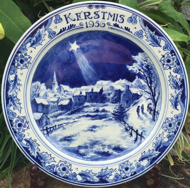 K&ouml;nnte beinhalten: Blaue und wei&szlig;e Keramikplatte mit einer Weihnachts-Szene, die ein Dorf unter einem sternenklaren Nachthimmel zeigt. Die Platte hat einen blauen und wei&szlig;en floralen Rand und den Text "KERSTMIS 1956" in der oberen Mitte.