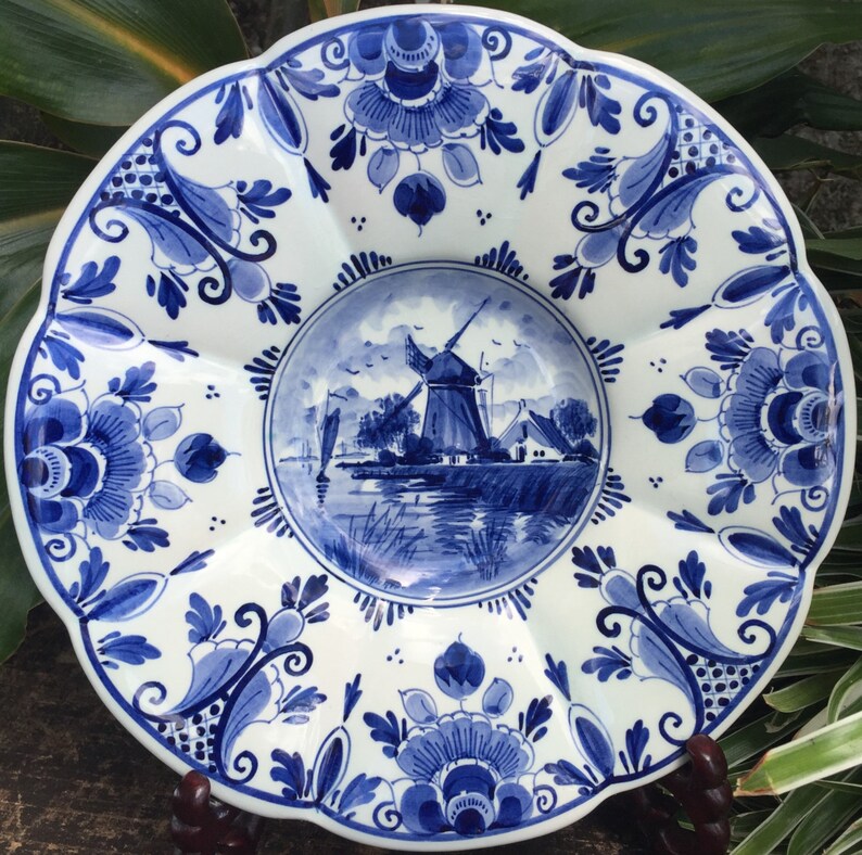 May include: A white ceramic plate with a blue and white windmill scene in the center. The plate has a scalloped edge and is decorated with blue and white floral designs.