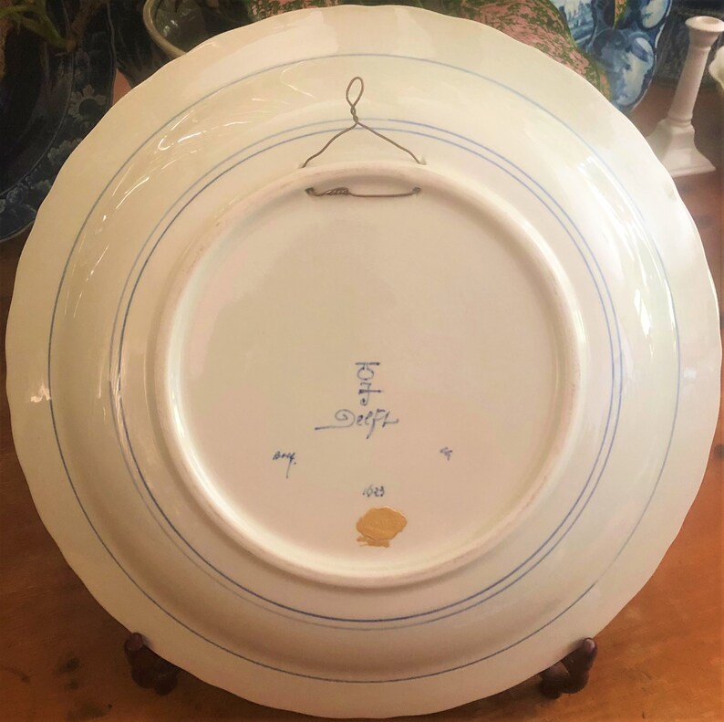 K&ouml;nnte beinhalten: Ein wei&szlig;er Keramikteller mit blauem Rand und einem goldenen Siegel. Auf der R&uuml;ckseite des Tellers befindet sich eine blaue Inschrift mit der Aufschrift "Kopf Delft" und eine goldene Zahl "1833".