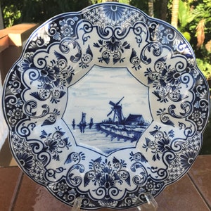 K&ouml;nnte beinhalten: Ein wei&szlig;er Teller aus Keramik mit einem blauen und wei&szlig;en Blumendesign. Die Mitte des Tellers zeigt eine blaue und wei&szlig;e Illustration einer Windm&uuml;hle und eines Kanals.