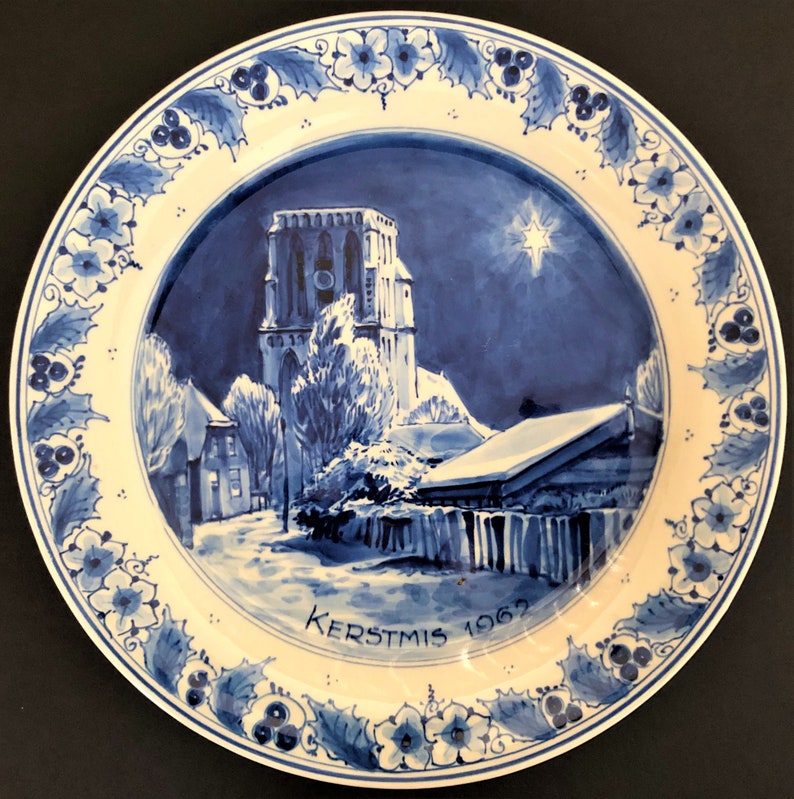 May include: A blue and white ceramic plate featuring a winter scene of a church and houses. The plate has a blue and white border with holly and berries. The text "KERSTMIS 1962" is written on the bottom of the plate.