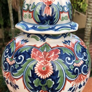 Może przedstawiać: Ozdobny ceramiczny słoik na imbir z pokrywką. Słoik jest kremowy i zdobiony kwiatowym wzorem w kolorach czerwonym, niebieskim i zielonym. Słoik stoi na czarnej podstawie i nadaje się do dekoracji wnętrz.