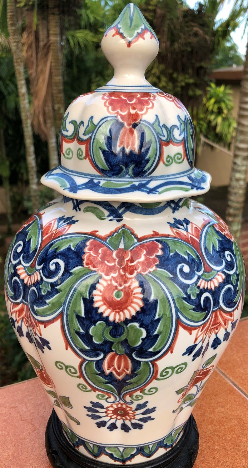 Może przedstawiać: Ozdobny ceramiczny słoik na imbir z pokrywką. Słoik jest kremowy i zdobiony kwiatowym wzorem w kolorach czerwonym, niebieskim i zielonym. Słoik stoi na czarnej podstawie i nadaje się do dekoracji wnętrz.