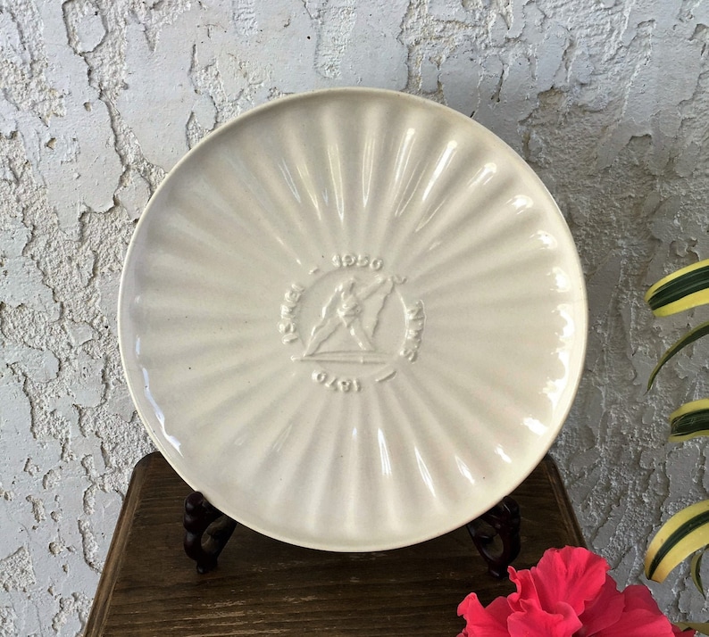 May include: A white ceramic plate with a raised design of a circle with the text "15th Annual" and "1950" above and "Olympics" below. The circle also has a stylized image of two figures holding a torch.