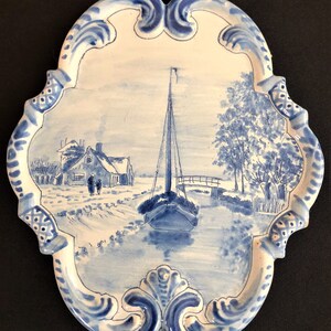 May include: A blue and white ceramic wall plaque with a winter landscape scene. The scene depicts a sailboat on a waterway, a house, and a bridge. The plaque has a decorative, ornate border and a string for hanging.