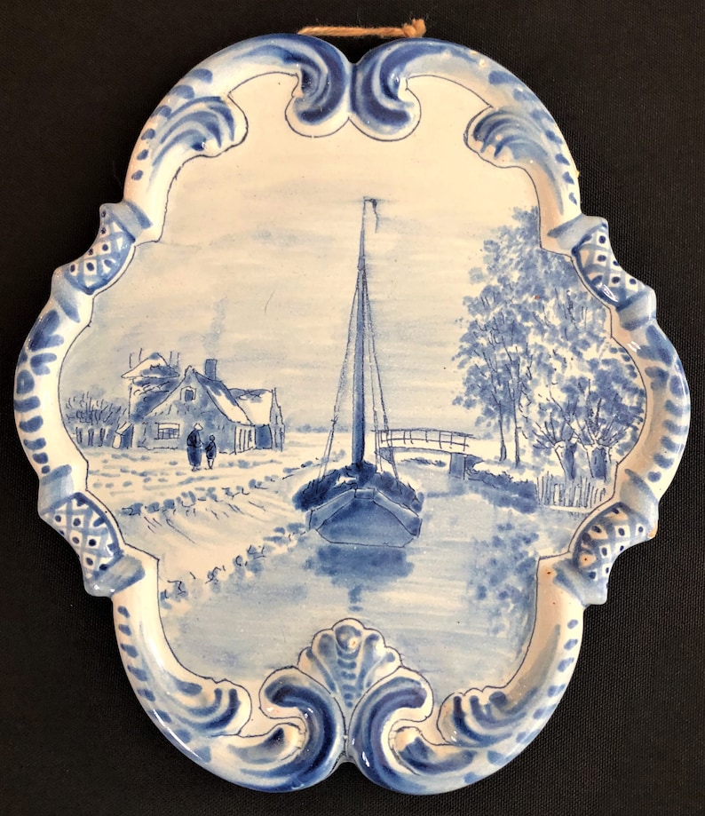 May include: A blue and white ceramic wall plaque with a winter landscape scene. The scene depicts a sailboat on a waterway, a house, and a bridge. The plaque has a decorative, ornate border and a string for hanging.