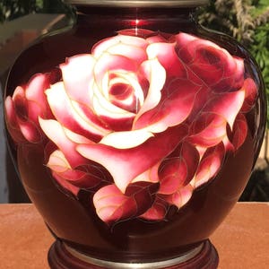 May include: A maroon-colored vase with a large, detailed rose design. The rose features white and red petals with gold accents. The vase sits on a dark wooden base and has a metallic rim.
