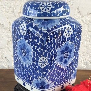 May include: A blue and white ceramic jar with a lid. The jar features a floral pattern with a square shape and a white base. The lid has a matching floral design. The jar is sitting on a black stand.