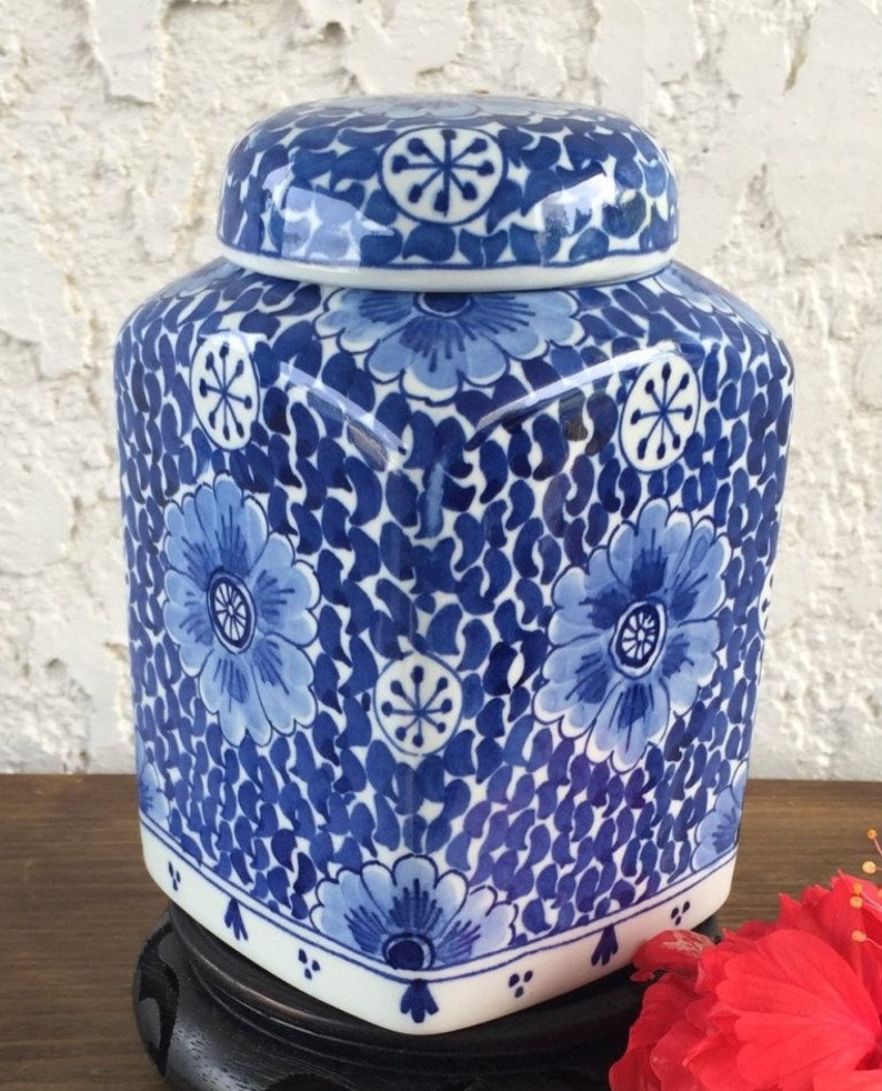 May include: A blue and white ceramic jar with a lid. The jar features a floral pattern with a square shape and a white base. The lid has a matching floral design. The jar is sitting on a black stand.