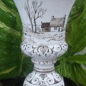 Puede incluir: Jarr&oacute;n de cer&aacute;mica blanca con parte superior acampanada y base de pedestal. El jarr&oacute;n presenta una escena de paisaje pintada a mano en tonos marrones y grises, que representa un entorno rural con una casa y &aacute;rboles. Adornos decorativos adornan la base y el borde.