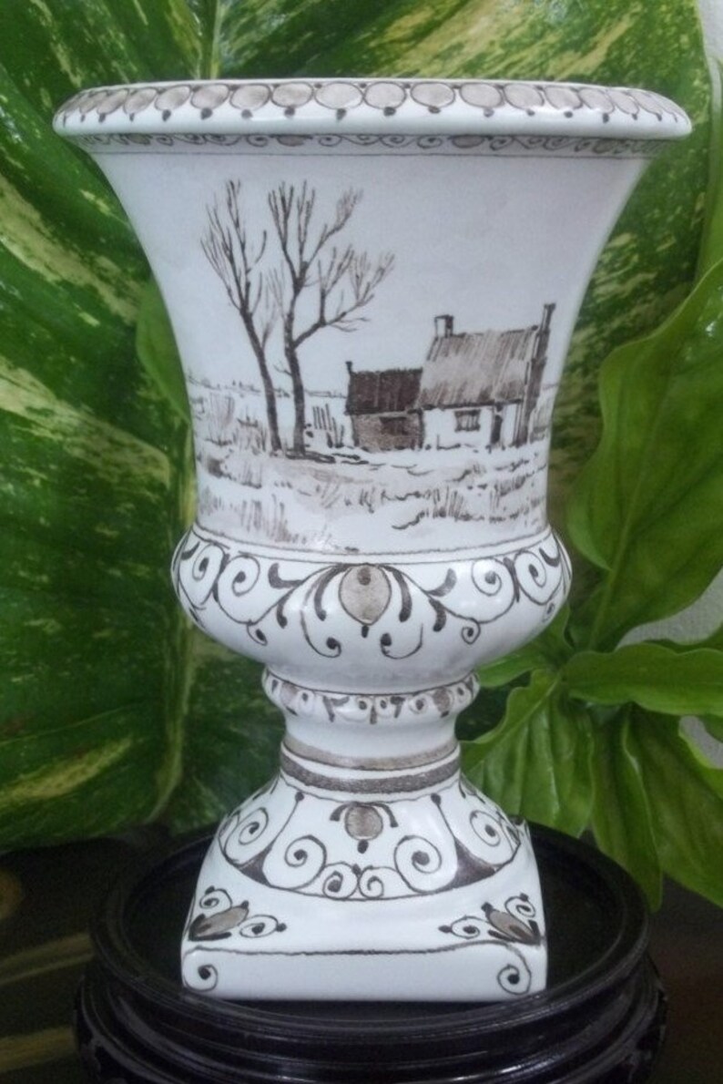 Puede incluir: Jarr&oacute;n de cer&aacute;mica blanca con parte superior acampanada y base de pedestal. El jarr&oacute;n presenta una escena de paisaje pintada a mano en tonos marrones y grises, que representa un entorno rural con una casa y &aacute;rboles. Adornos decorativos adornan la base y el borde.