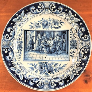 May include: A round, white and blue decorative plate with a detailed painting of a biblical scene. The plate features floral and scrollwork designs around the border. The painting in the center depicts figures in a room, with the text "LUCAS 2/V/23" below.