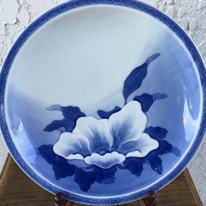 Op de afbeelding: Een wit en blauw keramisch bord met een bloemenontwerp. Het bord heeft een grote witte bloem met blauwe details en bladeren. De rand heeft een decoratieve blauwe rand. Het bord wordt op een houten standaard gepresenteerd.