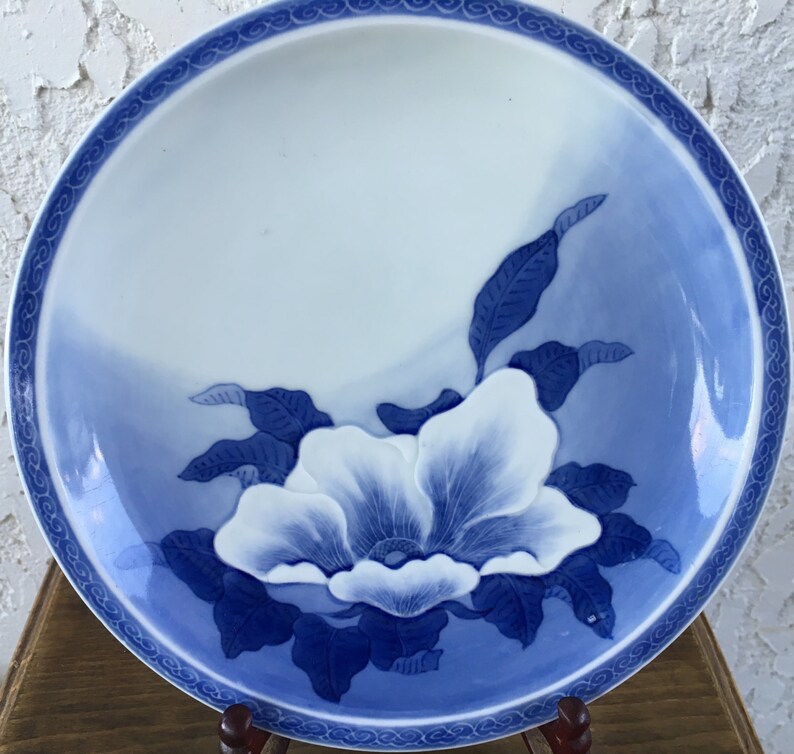 Op de afbeelding: Een wit en blauw keramisch bord met een bloemenontwerp. Het bord heeft een grote witte bloem met blauwe details en bladeren. De rand heeft een decoratieve blauwe rand. Het bord wordt op een houten standaard gepresenteerd.