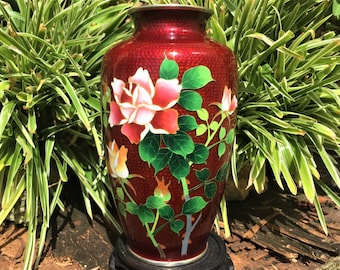 May include: A red cloisonné vase with a floral design. The vase features a textured surface and is adorned with pink and orange roses, green leaves, and stems. The vase is set against a backdrop of green foliage.