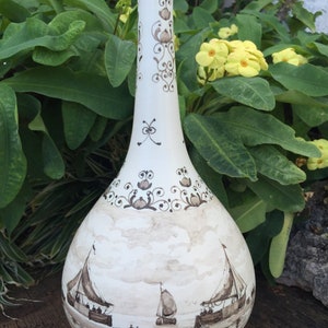 Puede incluir: Un jarr&oacute;n de cer&aacute;mica blanco con cuello largo y cuerpo bulboso. El jarr&oacute;n est&aacute; decorado con escenas n&aacute;uticas marrones de veleros y patrones florales decorativos. El jarr&oacute;n se exhibe sobre un fondo de follaje verde y flores amarillas.