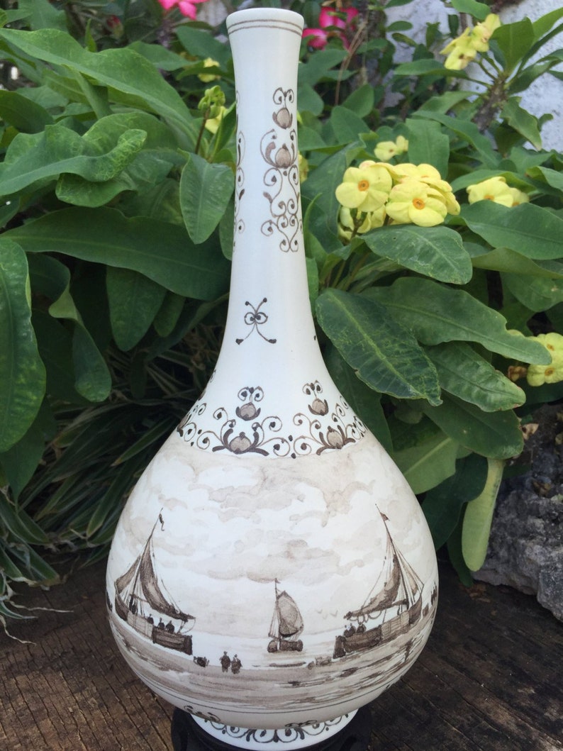Puede incluir: Un jarr&oacute;n de cer&aacute;mica blanco con cuello largo y cuerpo bulboso. El jarr&oacute;n est&aacute; decorado con escenas n&aacute;uticas marrones de veleros y patrones florales decorativos. El jarr&oacute;n se exhibe sobre un fondo de follaje verde y flores amarillas.