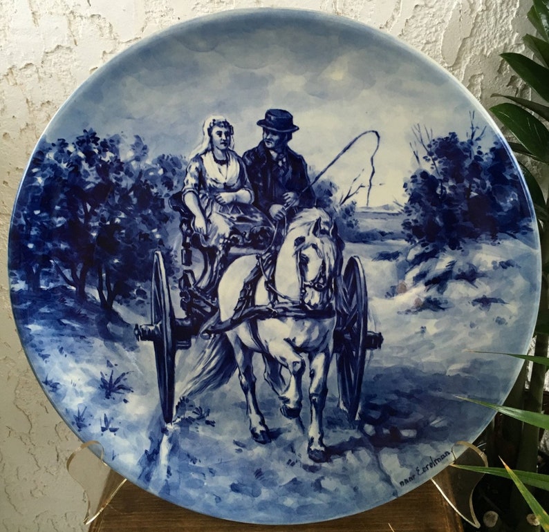 Pu&ograve; includere: Un piatto in ceramica blu e bianco con una scena di una coppia che viaggia in una carrozza trainata da un cavallo. Il piatto &egrave; decorato con un motivo floreale blu e bianco e il testo "naar een ontwerp" nell'angolo in basso a destra.