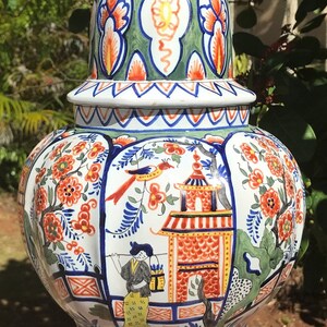 K&ouml;nnte beinhalten: Eine gro&szlig;e, wei&szlig;e Keramikvase mit Deckel und einem Vogelaufsatz. Die Vase ist mit einem bunten Blumenmuster und einer Szene mit einer Pagode und einer Frau in traditioneller Kleidung verziert. Die Vase ist ein traditionelles niederl&auml;ndisches Delftware-Design.