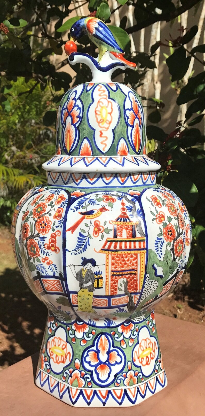K&ouml;nnte beinhalten: Eine gro&szlig;e, wei&szlig;e Keramikvase mit Deckel und einem Vogelaufsatz. Die Vase ist mit einem bunten Blumenmuster und einer Szene mit einer Pagode und einer Frau in traditioneller Kleidung verziert. Die Vase ist ein traditionelles niederl&auml;ndisches Delftware-Design.