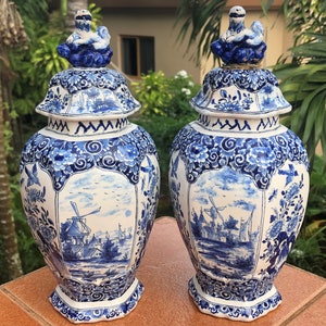 May include: A pair of blue and white ceramic vases with lids. The vases are decorated with a landscape scene including windmills and floral motifs. Each lid is adorned with a small figurine. The vases are shown outside.