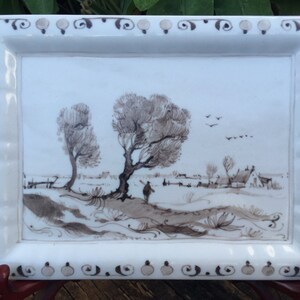 Puede incluir: Bandeja rectangular de cer&aacute;mica blanca con una escena de paisaje en tonos sepia. La escena representa &aacute;rboles, una figura y un pueblo. El borde de la bandeja presenta un patr&oacute;n decorativo de espirales y puntos.