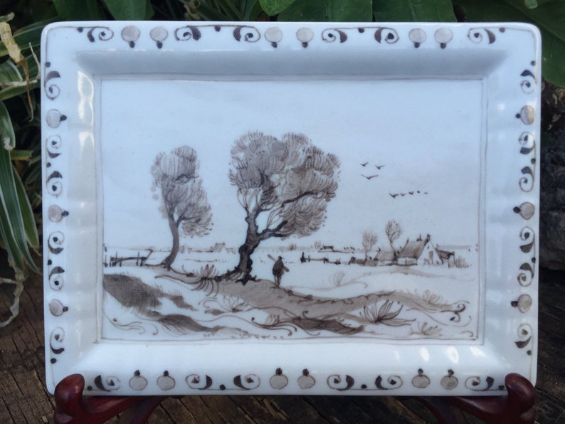 Puede incluir: Bandeja rectangular de cer&aacute;mica blanca con una escena de paisaje en tonos sepia. La escena representa &aacute;rboles, una figura y un pueblo. El borde de la bandeja presenta un patr&oacute;n decorativo de espirales y puntos.