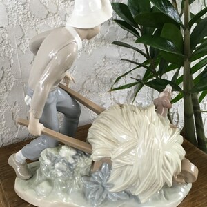 Puede incluir: Figura de porcelana que representa a una persona empujando una carretilla llena de heno. La figura lleva un sombrero blanco, un su&eacute;ter gris claro y pantalones azules. El heno es de color blanco cremoso y la base tiene un dise&ntilde;o floral verde claro y azul.