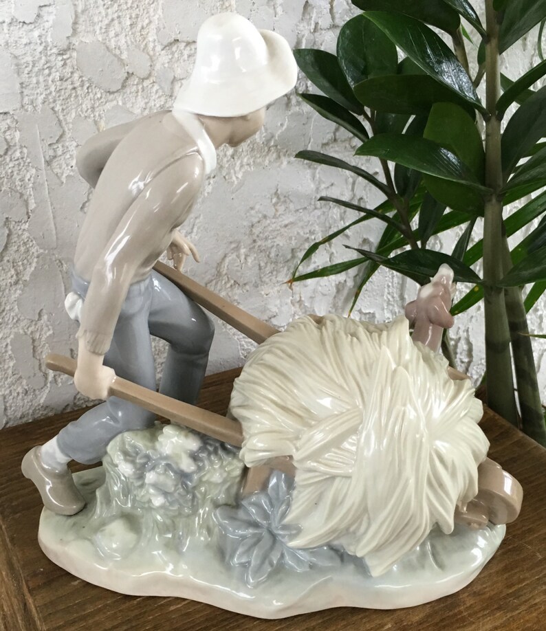 Puede incluir: Figura de porcelana que representa a una persona empujando una carretilla llena de heno. La figura lleva un sombrero blanco, un su&eacute;ter gris claro y pantalones azules. El heno es de color blanco cremoso y la base tiene un dise&ntilde;o floral verde claro y azul.