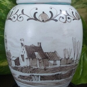 May include: A white ceramic jar with a lid, featuring a hand-painted landscape scene in shades of brown and gray. The jar has decorative floral patterns around the top and base, and a light blue band near the lid.