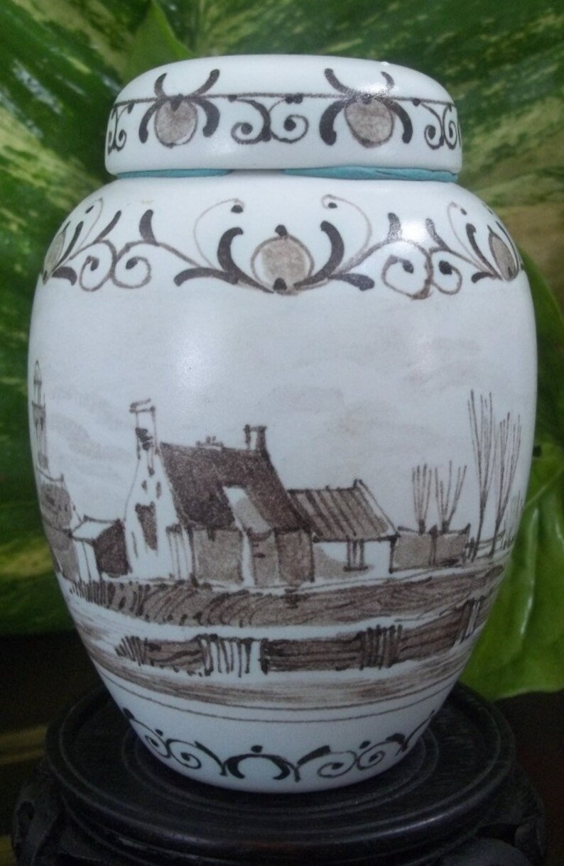 May include: A white ceramic jar with a lid, featuring a hand-painted landscape scene in shades of brown and gray. The jar has decorative floral patterns around the top and base, and a light blue band near the lid.