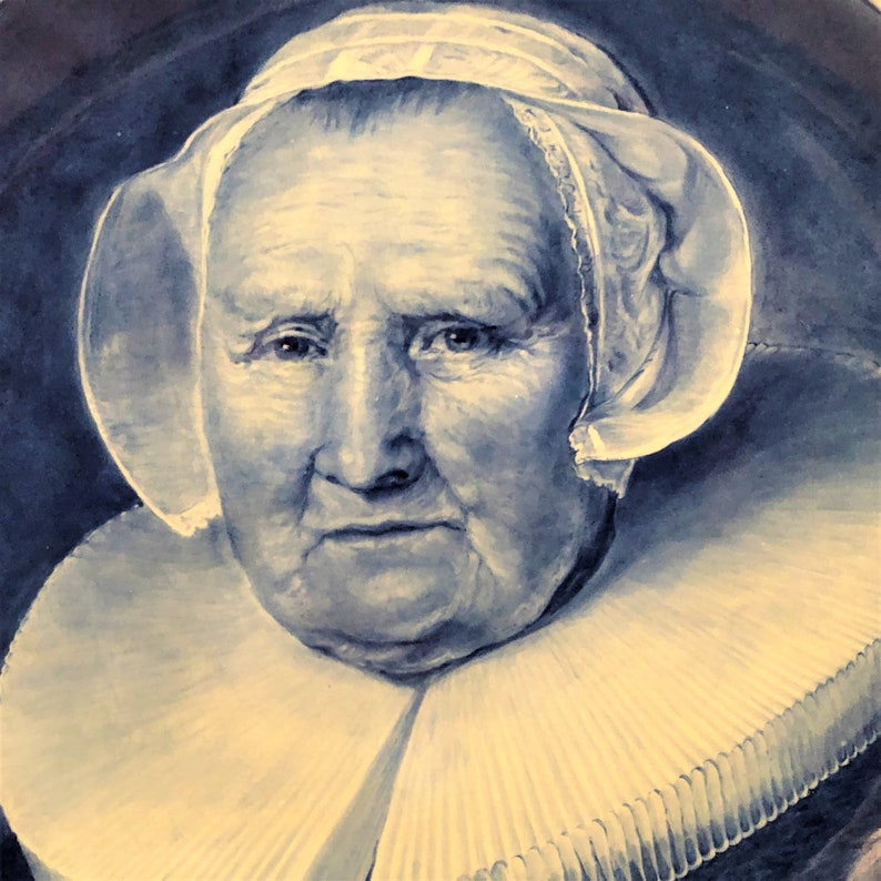 May include: A close-up portrait of an older woman wearing a white lace cap and a large white ruffled collar. The background is a dark blue.