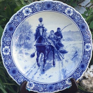 Pu&ograve; includere: Un piatto in maiolica di Delft blu e bianco con una scena invernale di una slitta trainata da un cavallo. Il piatto ha un bordo decorativo di fiori e volute blu e bianche.