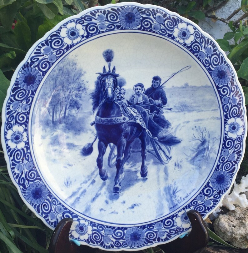 Pu&ograve; includere: Un piatto in maiolica di Delft blu e bianco con una scena invernale di una slitta trainata da un cavallo. Il piatto ha un bordo decorativo di fiori e volute blu e bianche.