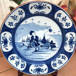 Puede incluir: Un plato de cer&aacute;mica azul y blanco con un dise&ntilde;o pintado a mano de una persona sentada en un banco con un jarr&oacute;n de flores. El plato tiene un borde floral azul y blanco.