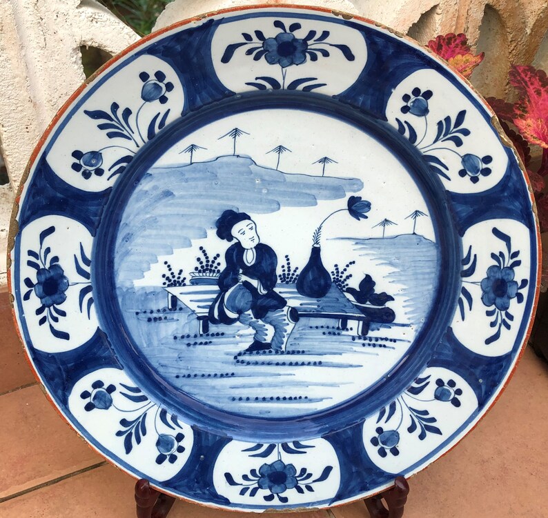 Puede incluir: Un plato de cer&aacute;mica azul y blanco con un dise&ntilde;o pintado a mano de una persona sentada en un banco con un jarr&oacute;n de flores. El plato tiene un borde floral azul y blanco.