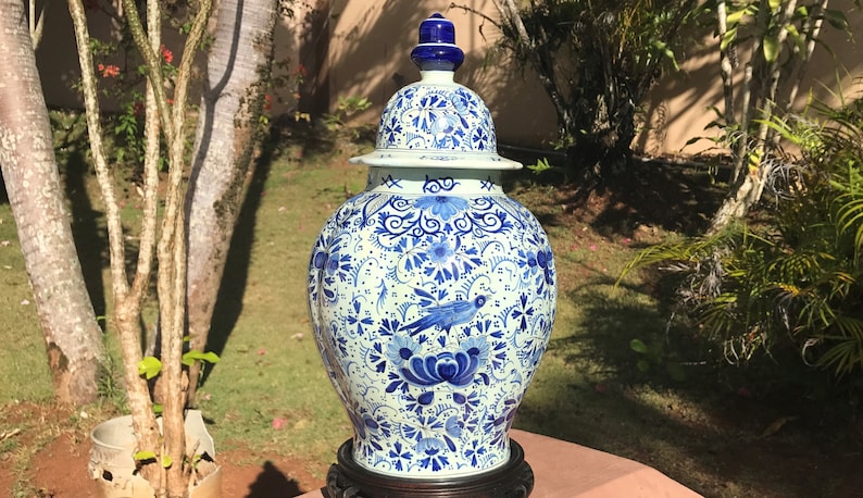 May include: A large, ornate blue and white ceramic jar with a lid. The jar features a detailed floral and bird pattern. It is displayed on a dark wooden stand, set against a backdrop of greenery.