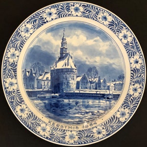 May include: A blue and white ceramic plate featuring a winter scene of a Dutch town. The plate has a white border with a blue floral design. The text "KERSTMIS 1964" is written on the bottom of the plate.