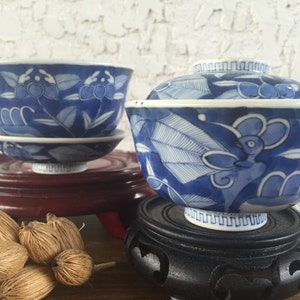 May include: Two blue and white porcelain bowls with floral designs. One bowl has a lid. The bowls are on decorative wooden stands. The bowls are decorated with blue floral patterns and are a classic example of Asian ceramics.