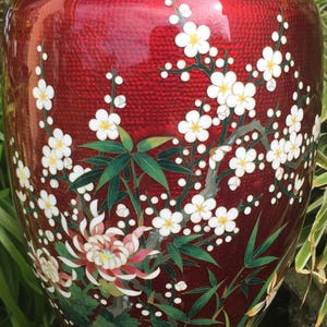 Puede incluir: Un jarr&oacute;n de cloisonn&eacute; rojo con un dise&ntilde;o floral. El jarr&oacute;n est&aacute; decorado con flores blancas, hojas verdes y detalles dorados. El jarr&oacute;n est&aacute; sobre una superficie de madera.