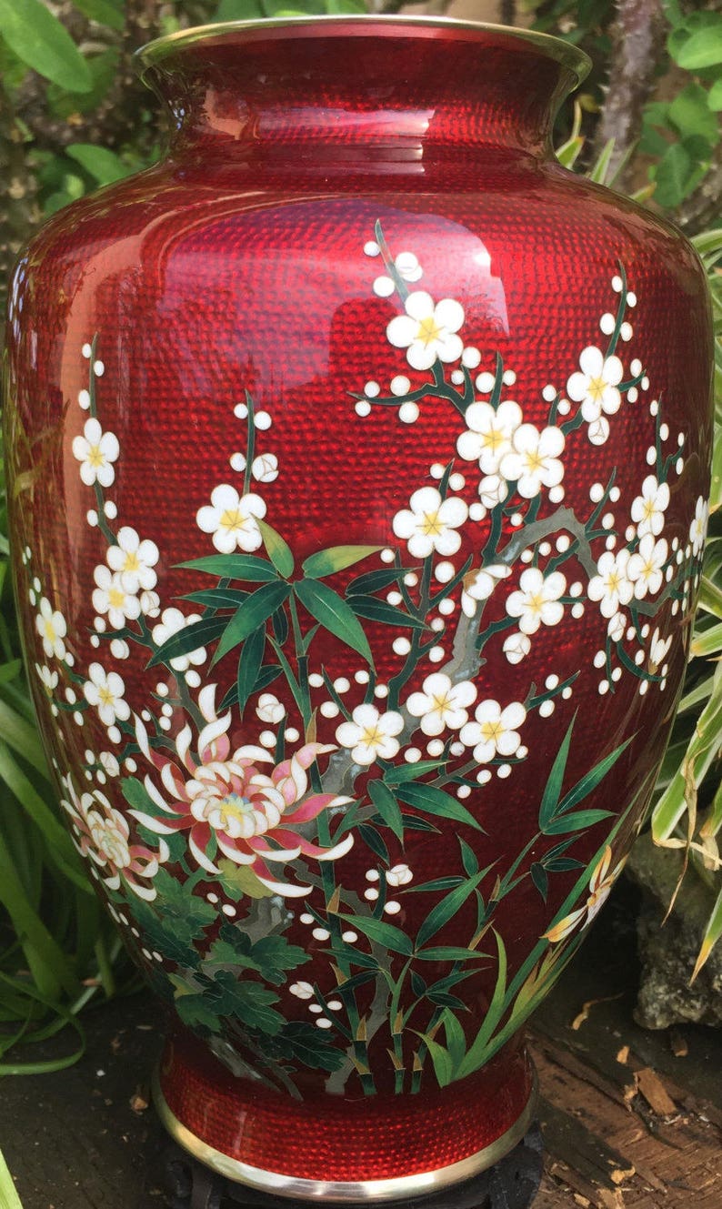 Puede incluir: Un jarr&oacute;n de cloisonn&eacute; rojo con un dise&ntilde;o floral. El jarr&oacute;n est&aacute; decorado con flores blancas, hojas verdes y detalles dorados. El jarr&oacute;n est&aacute; sobre una superficie de madera.