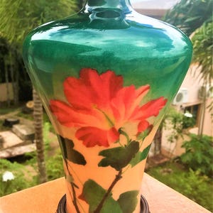 Puede incluir: Un jarr&oacute;n de cloisonn&eacute; verde con un dise&ntilde;o floral que presenta una flor de hibisco roja y hojas verdes. El jarr&oacute;n tiene un borde dorado y se asienta sobre una base de madera.