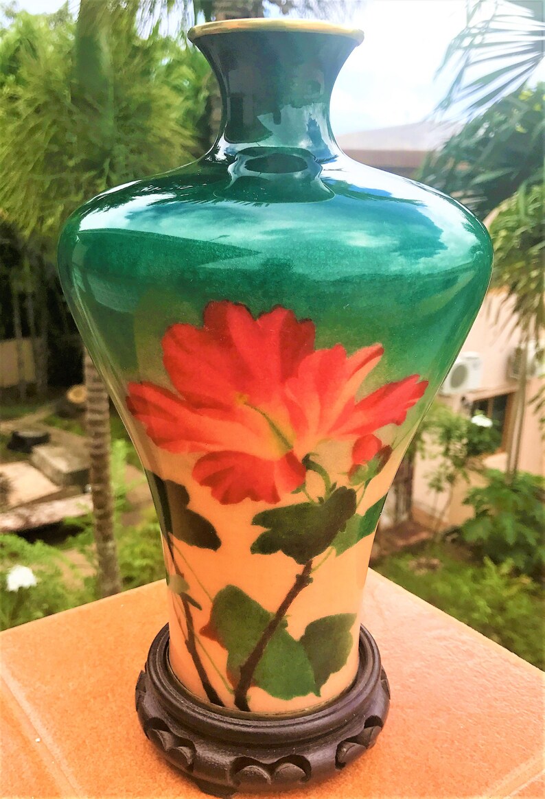 Puede incluir: Un jarr&oacute;n de cloisonn&eacute; verde con un dise&ntilde;o floral que presenta una flor de hibisco roja y hojas verdes. El jarr&oacute;n tiene un borde dorado y se asienta sobre una base de madera.