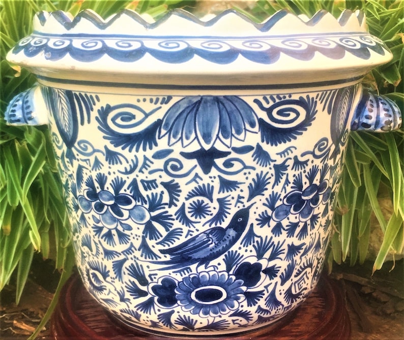May include: A large, round ceramic planter with a white base and intricate blue floral and bird designs. The planter has two handles and a scalloped rim. The design is reminiscent of Delftware pottery.