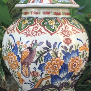 May include: A white ceramic jar with a lid, featuring a colorful floral and bird design. The jar has a bulbous shape and is decorated with orange, blue, and yellow flowers, and a bird perched on a branch. The lid is topped with a blue finial.