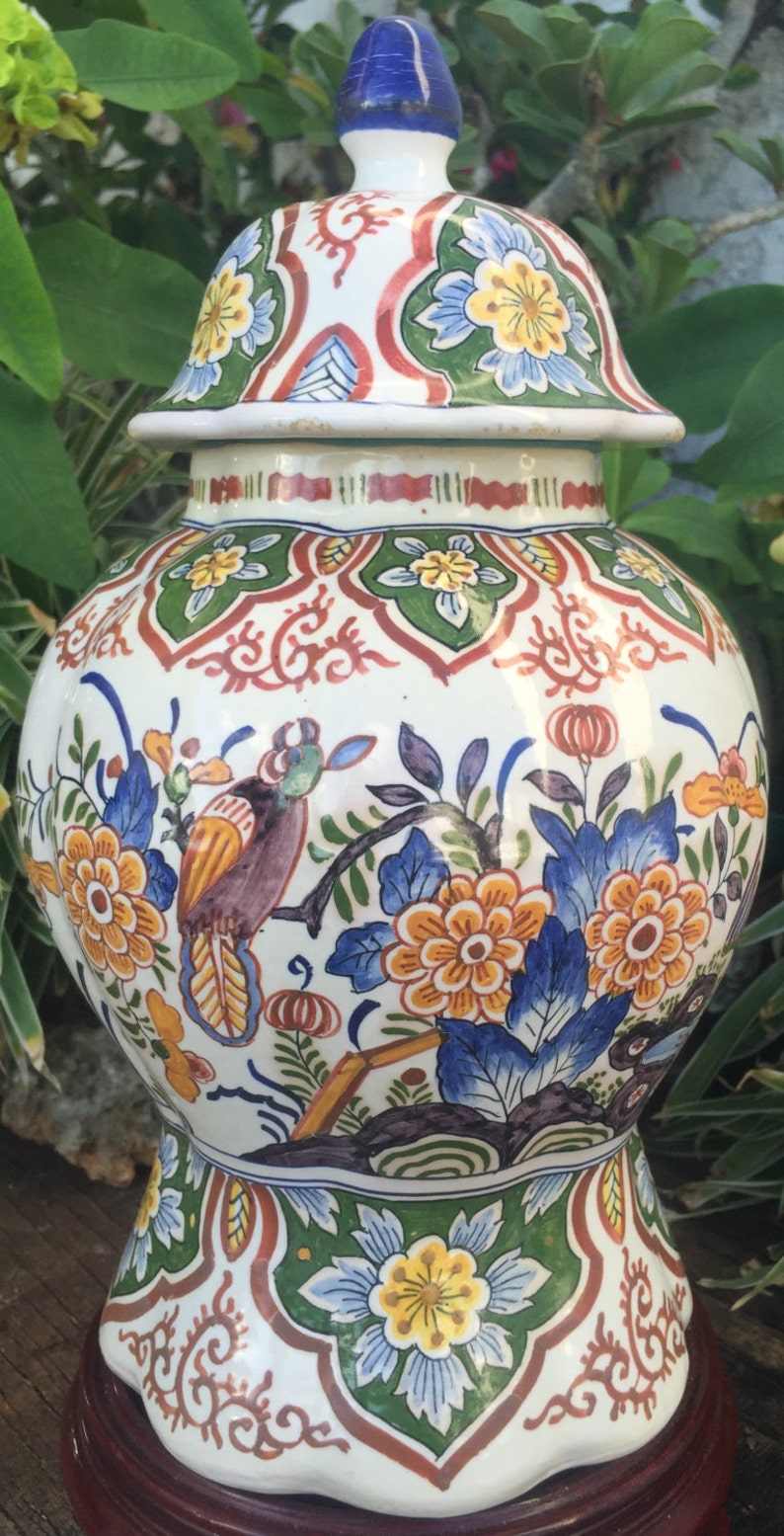 May include: A white ceramic jar with a lid, featuring a colorful floral and bird design. The jar has a bulbous shape and is decorated with orange, blue, and yellow flowers, and a bird perched on a branch. The lid is topped with a blue finial.